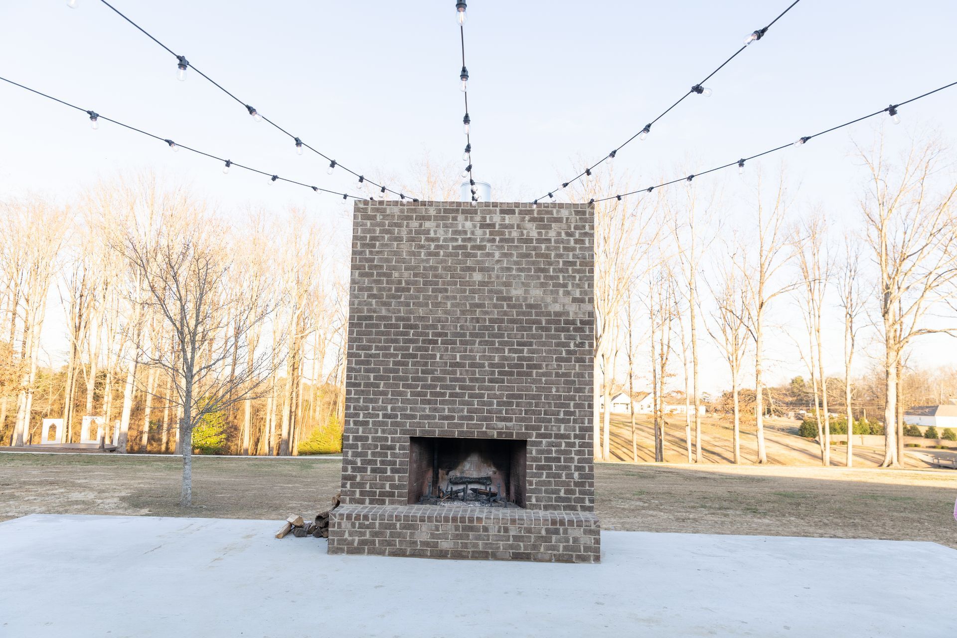 Brick outdoor fireplace with string lights overhead on a patio with bare trees in the background.