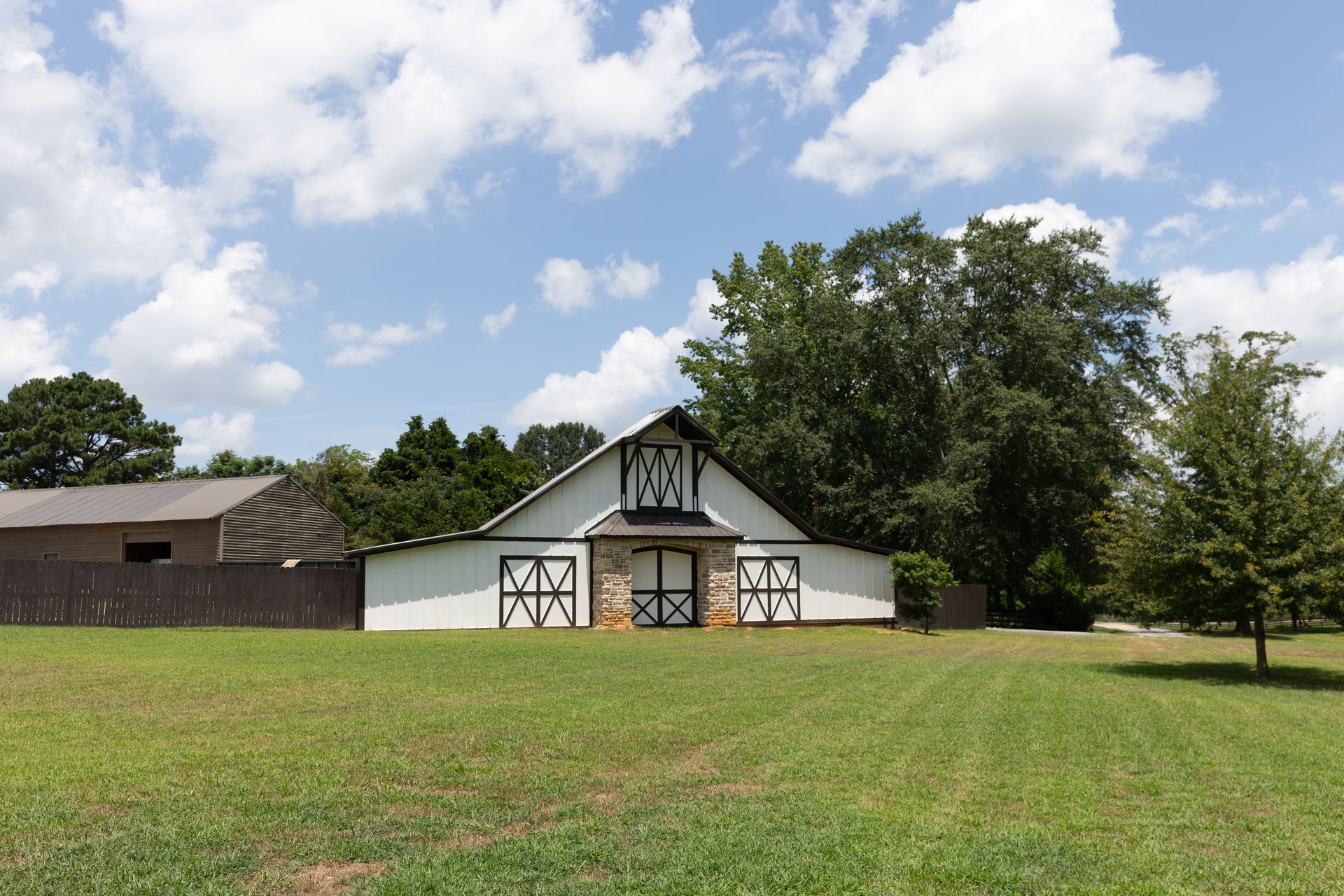 White barn with black trim and doors, stone facade, on a grassy field under a blue sky with clouds.