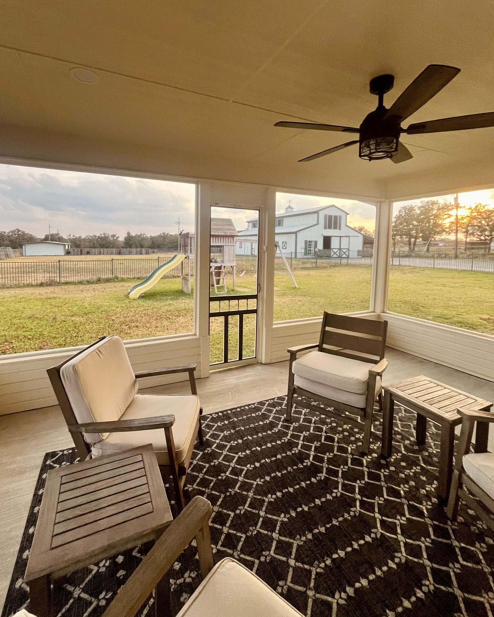 Covered patio with wicker chairs, patterned rug, and ceiling fan overlooking a grassy backyard and neighboring houses