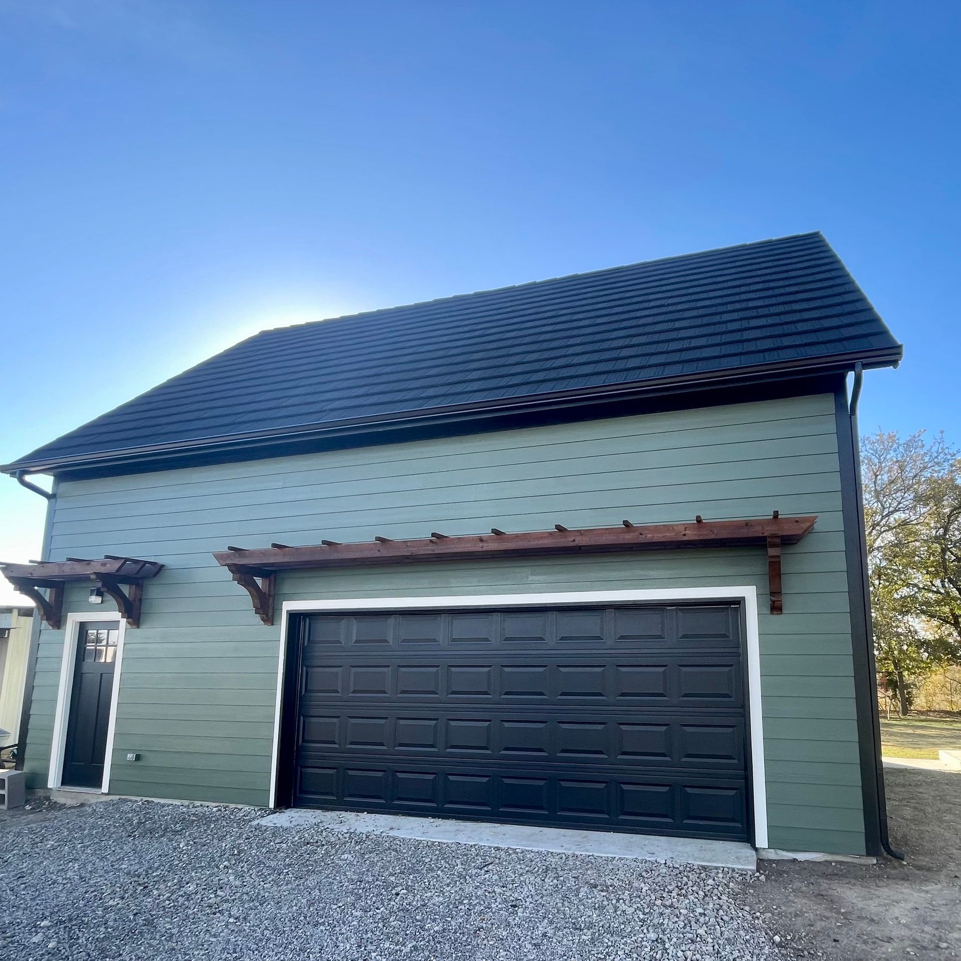 Small two-story house with dark roof, black garage door, and light green siding on a gravel lot.