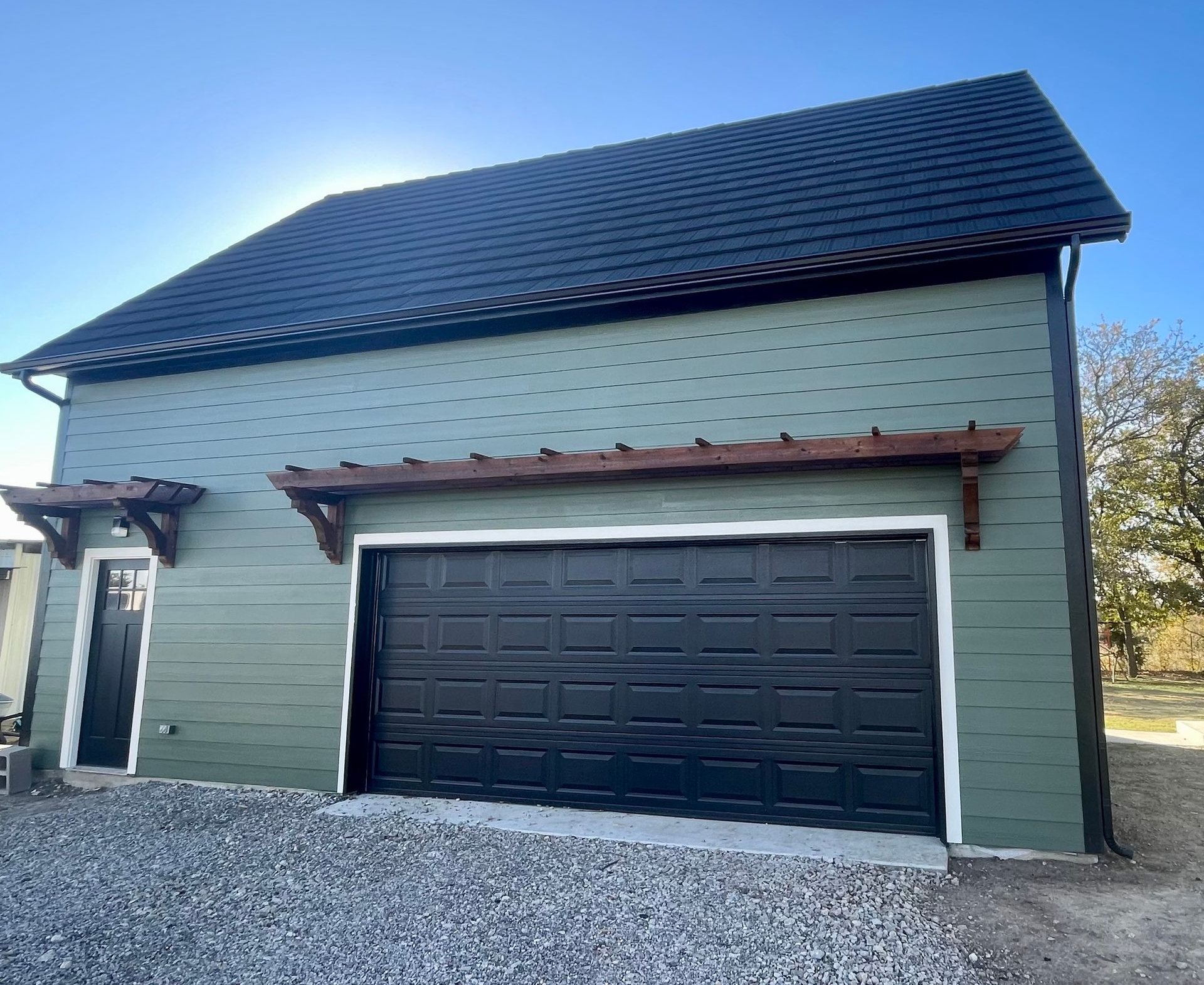 Modern two-story garage with dark roof, pale green siding, and black double garage door on gravel driveway