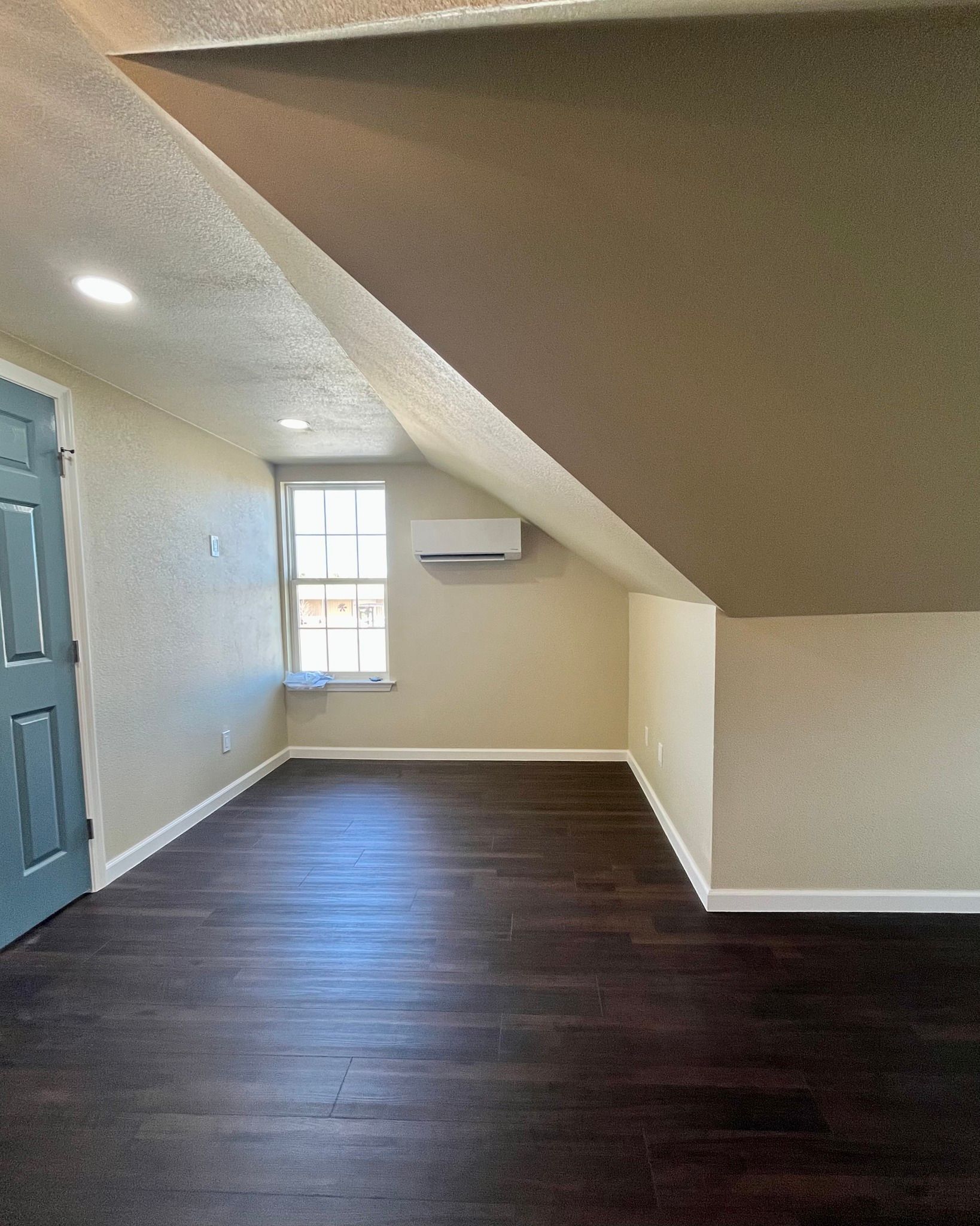 Empty attic-style room with dark wood floor, beige walls, blue door, and small window with wall AC unit