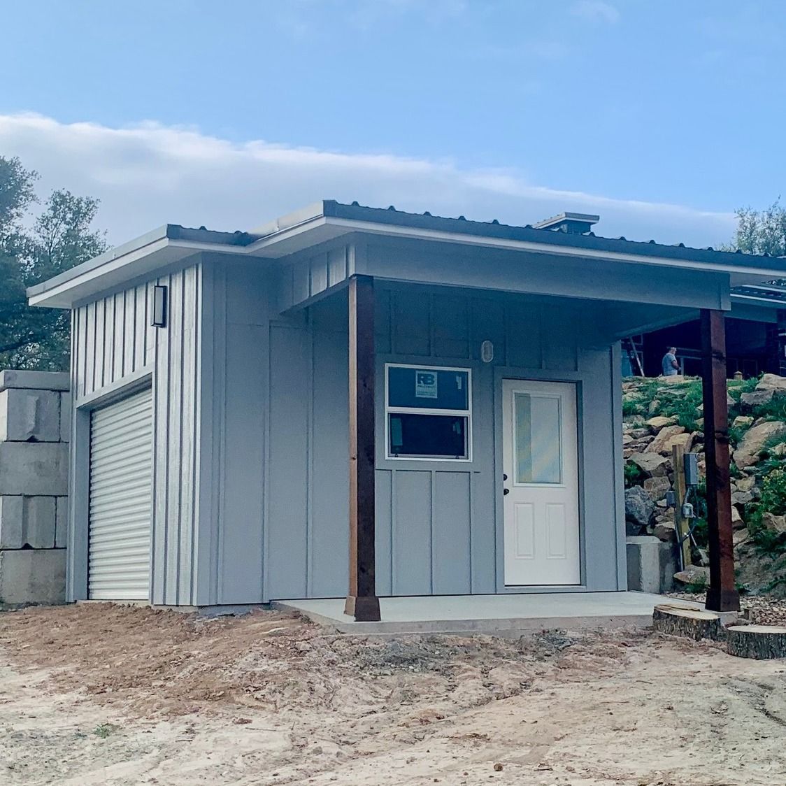 Small gray shed-style building with a porch, white door, and garage door in a dirt yard.