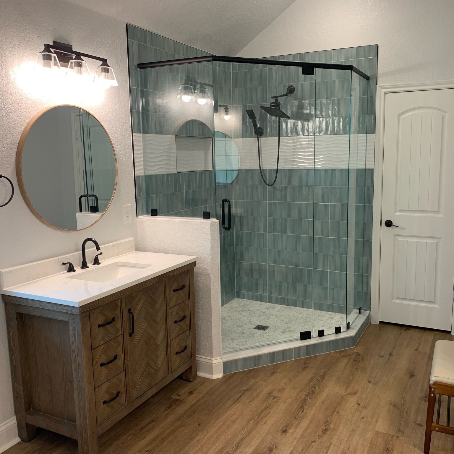 Modern bathroom with wood vanity, round mirror, and glass walk-in shower with teal tile.