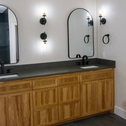 Modern bathroom vanity with wood cabinets, black countertop, arched mirror, and wall sconces.