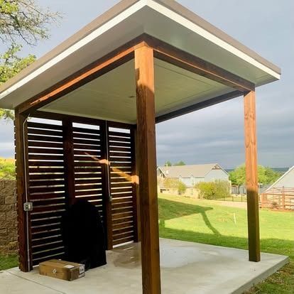 Modern outdoor pavilion with wooden posts, slatted wall, and concrete floor overlooking a grassy yard