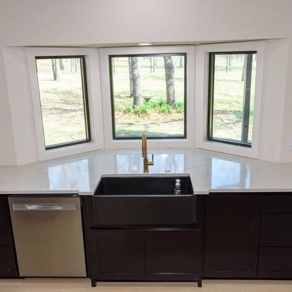 Modern kitchen sink beneath bay windows, with dark cabinets and a stainless dishwasher.