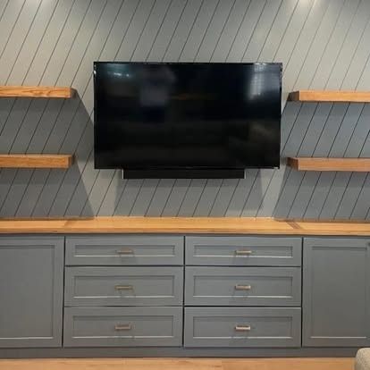Wall-mounted black TV above gray cabinet with drawers and shelves on a gray paneled wall.