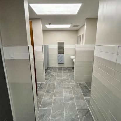 Clean public restroom with gray tile floors, white stalls, sinks, and bright ceiling lights.