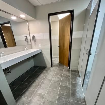 Modern public restroom with gray tile floor, white sink counter, mirror, and open wooden door to a stall