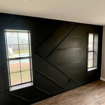 Black interior wall with two tall windows and diagonal trim, showing a view outside.