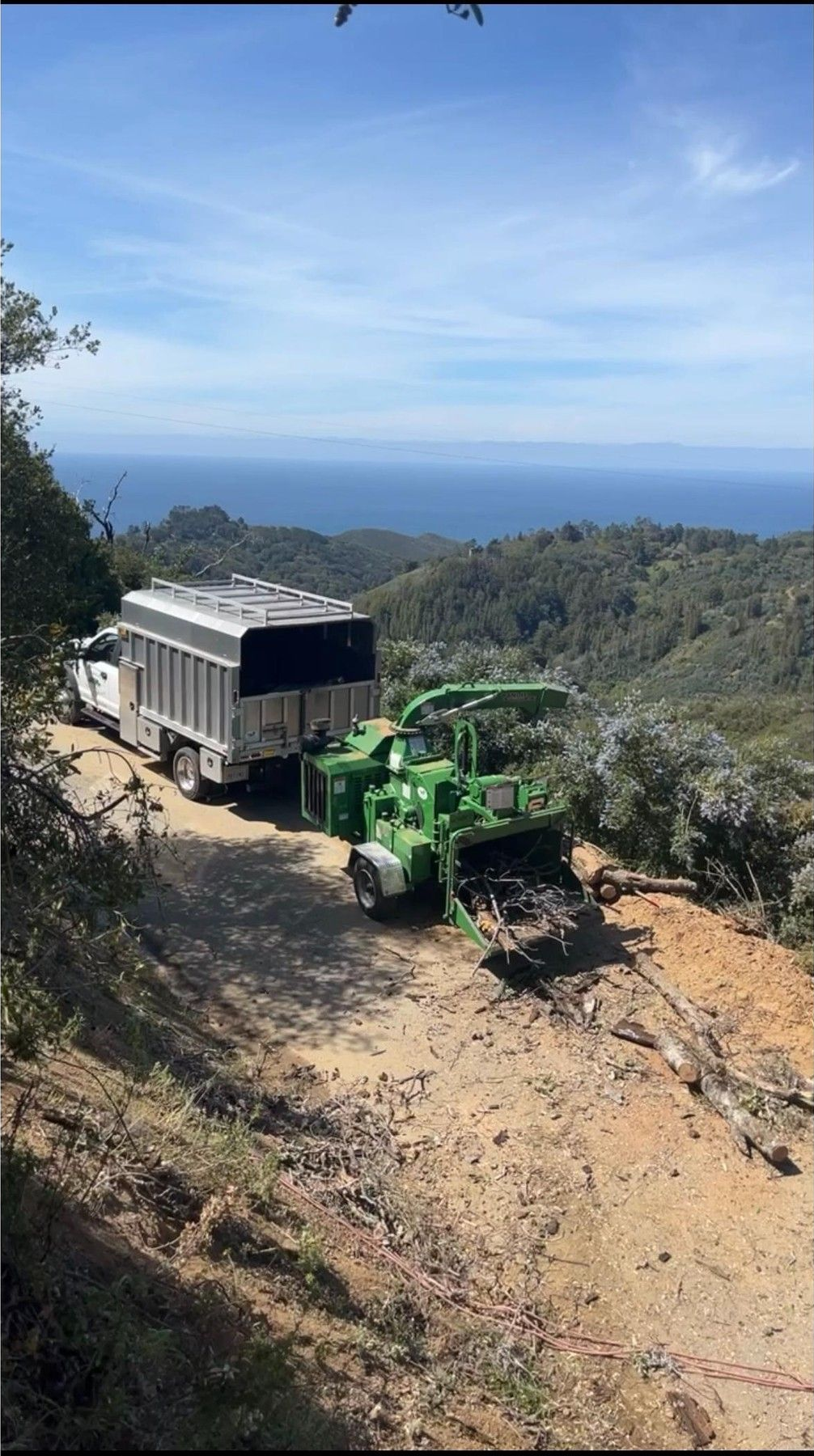 Truck and wood chipper on a hillside path overlooking the ocean. Green machine, white truck.