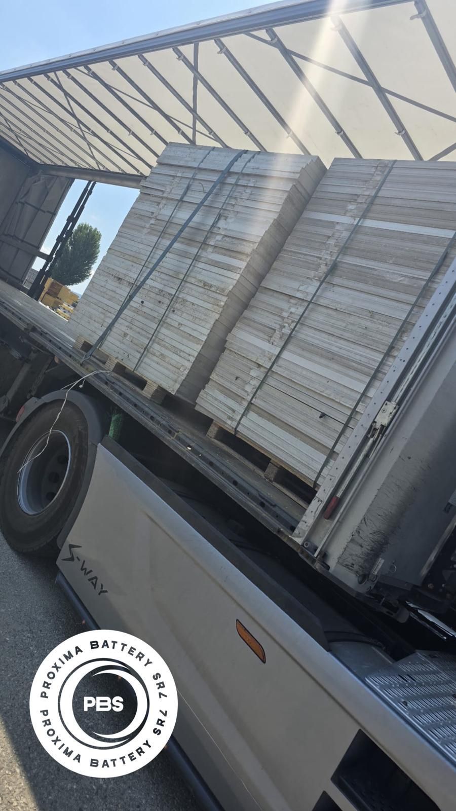 Un rimorchio di un camion carico di materiali rettangolari bianchi impilati, con sovrapposto il logo circolare 
