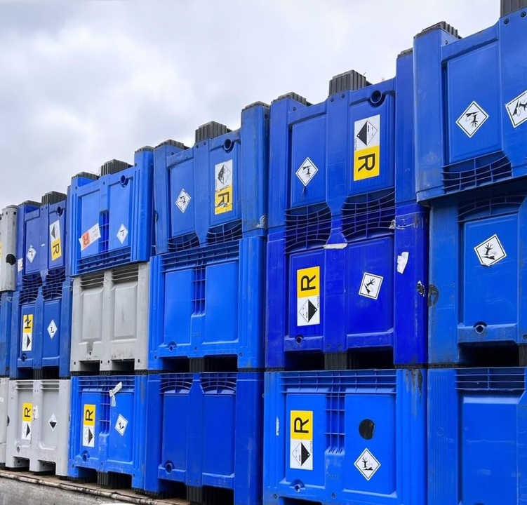 Pile di container industriali blu, contrassegnati da etichette di pericolo, posizionati all'aperto contro un cielo nuvoloso.