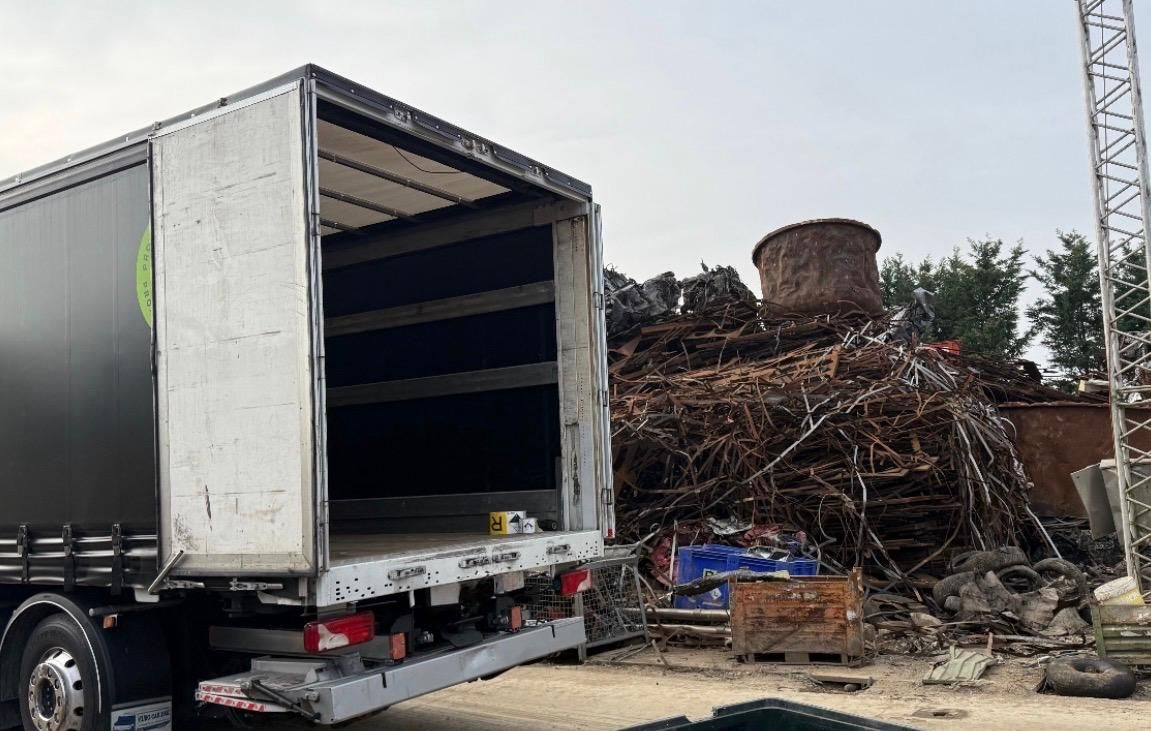 Un grande container nero a cielo aperto si trova di fronte a un semirimorchio vuoto parcheggiato presso un impianto di riciclaggio della Proxima Battery SRL.