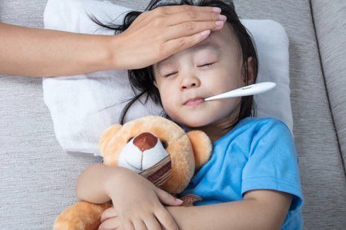 Child lying on couch with thermometer, holding a teddy bear, while an adult checks their forehead