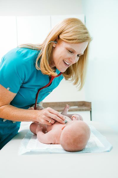 Nurse in teal uniform examining a baby on a changing table in a bright clinic room