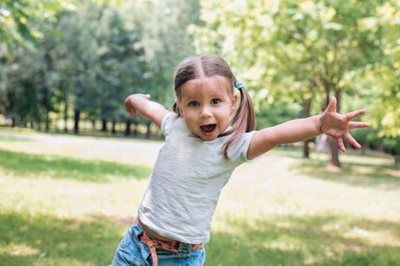 Child outdoors in a park, smiling with arms outstretched in a playful pose.