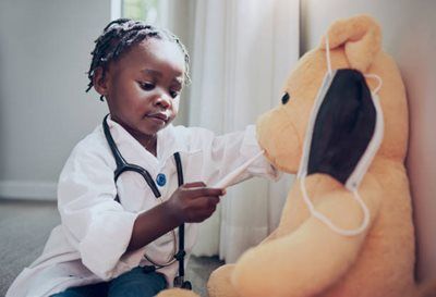Child in a white coat checks a teddy bear with a stethoscope in a bright room.