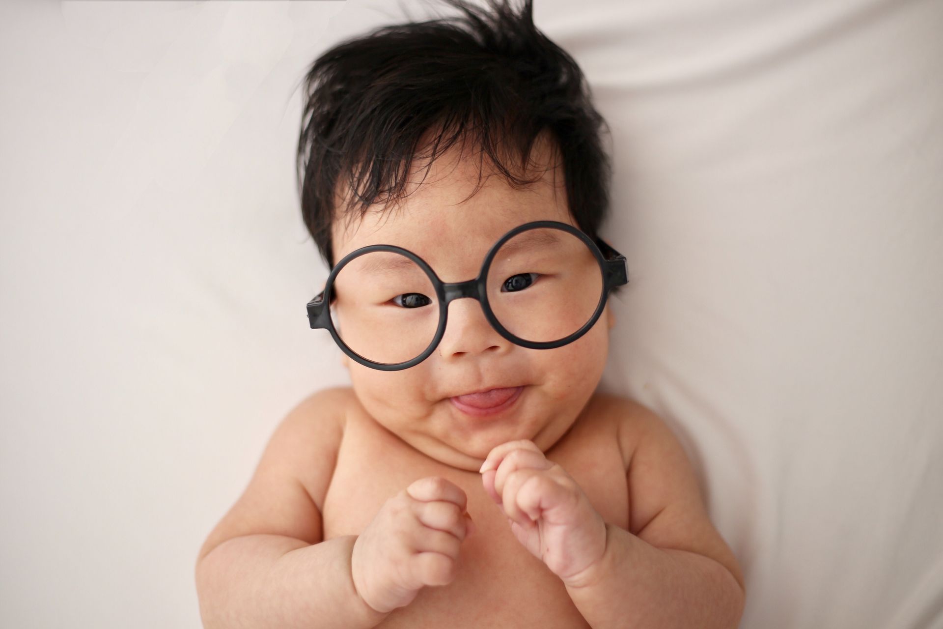 Smiling baby wearing oversized round black glasses, lying on a white blanket.