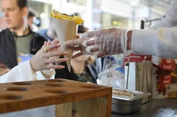 Person, die an einem Imbissstand eine Tüte Pommes aus der behandschuhten Hand eines Verkäufers erhält.