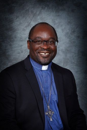 Smiling man in a dark suit and blue clerical collar, wearing a cross necklace, against a gray backdrop