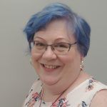 Smiling person with short blue hair, glasses, and a pink top against a light wall.