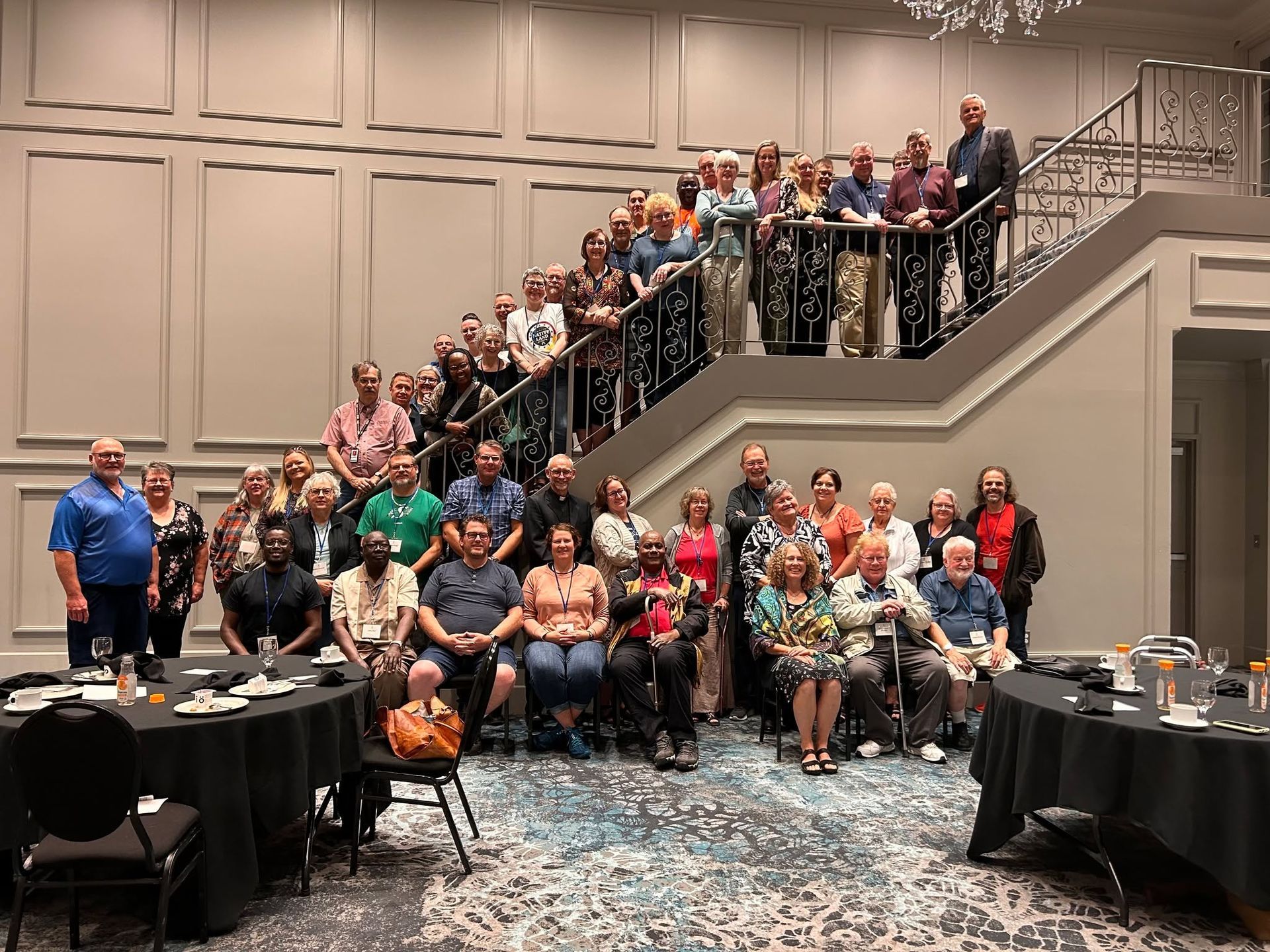 Large group posing on a staircase in a banquet hall, with tables and chairs in the foreground.
