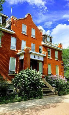 Red brick townhouse with white-trimmed windows, stone steps, and lush shrubs under a blue sky