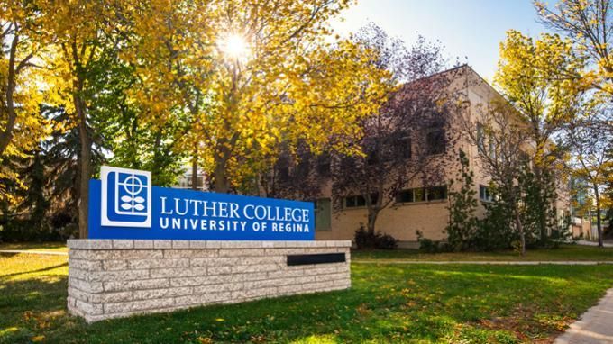 Luther College University of Regina sign beside a sunlit campus building and autumn trees