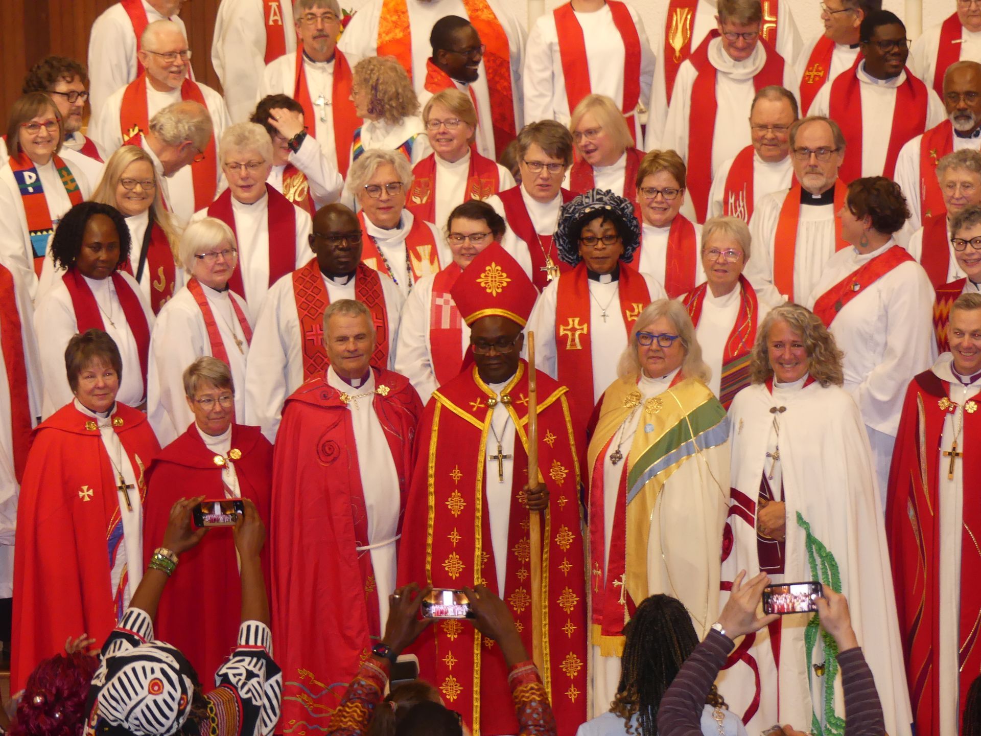 Clergy in red and white robes standing together in a church, with a central bishop in gold vestments