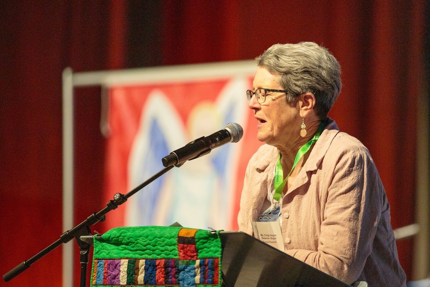 Woman speaking at a podium with a microphone, red curtain backdrop and colorful cloth draped over the lectern.