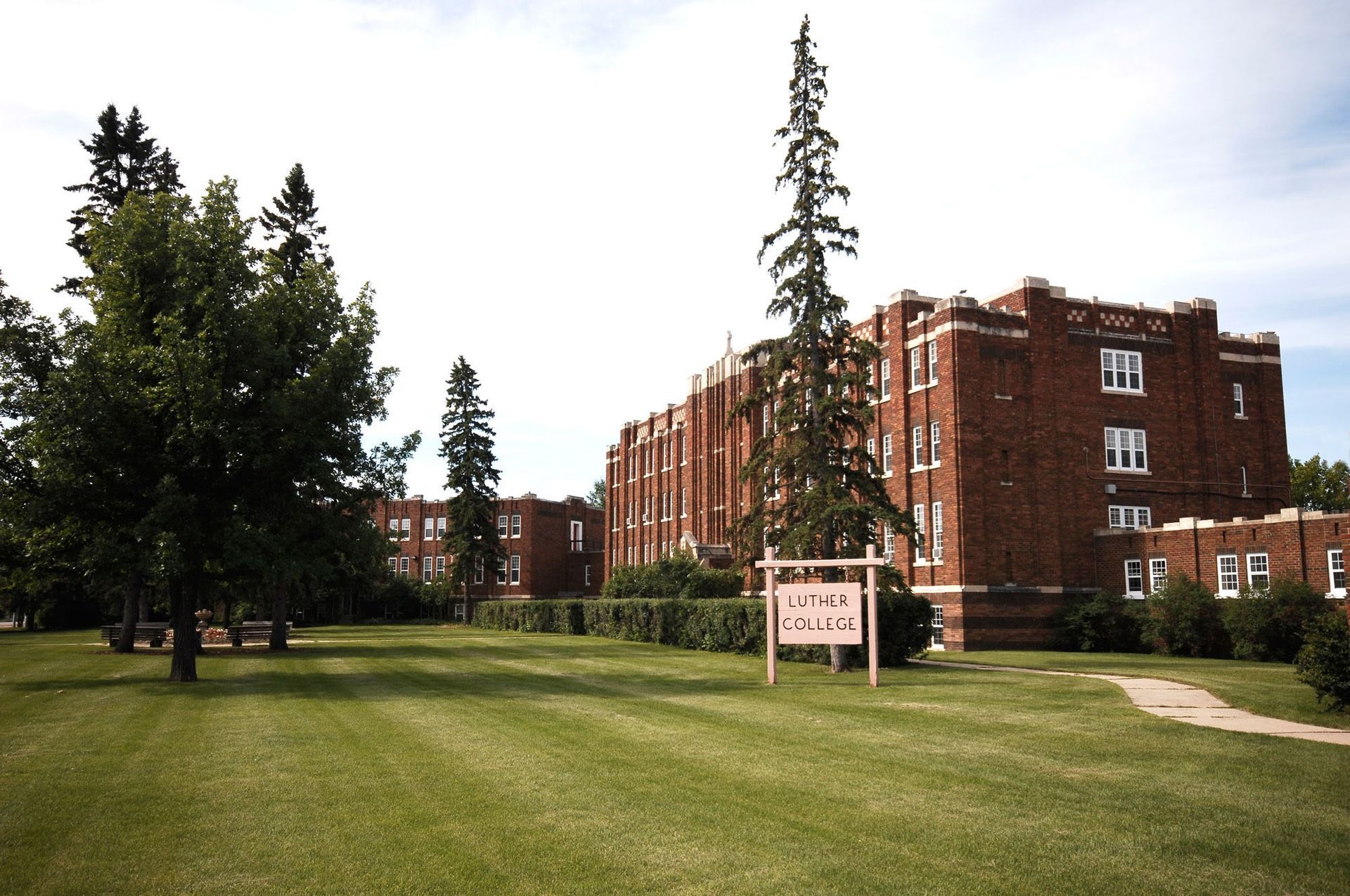 Brick campus building beside a green lawn with trees and a sign under a bright sky