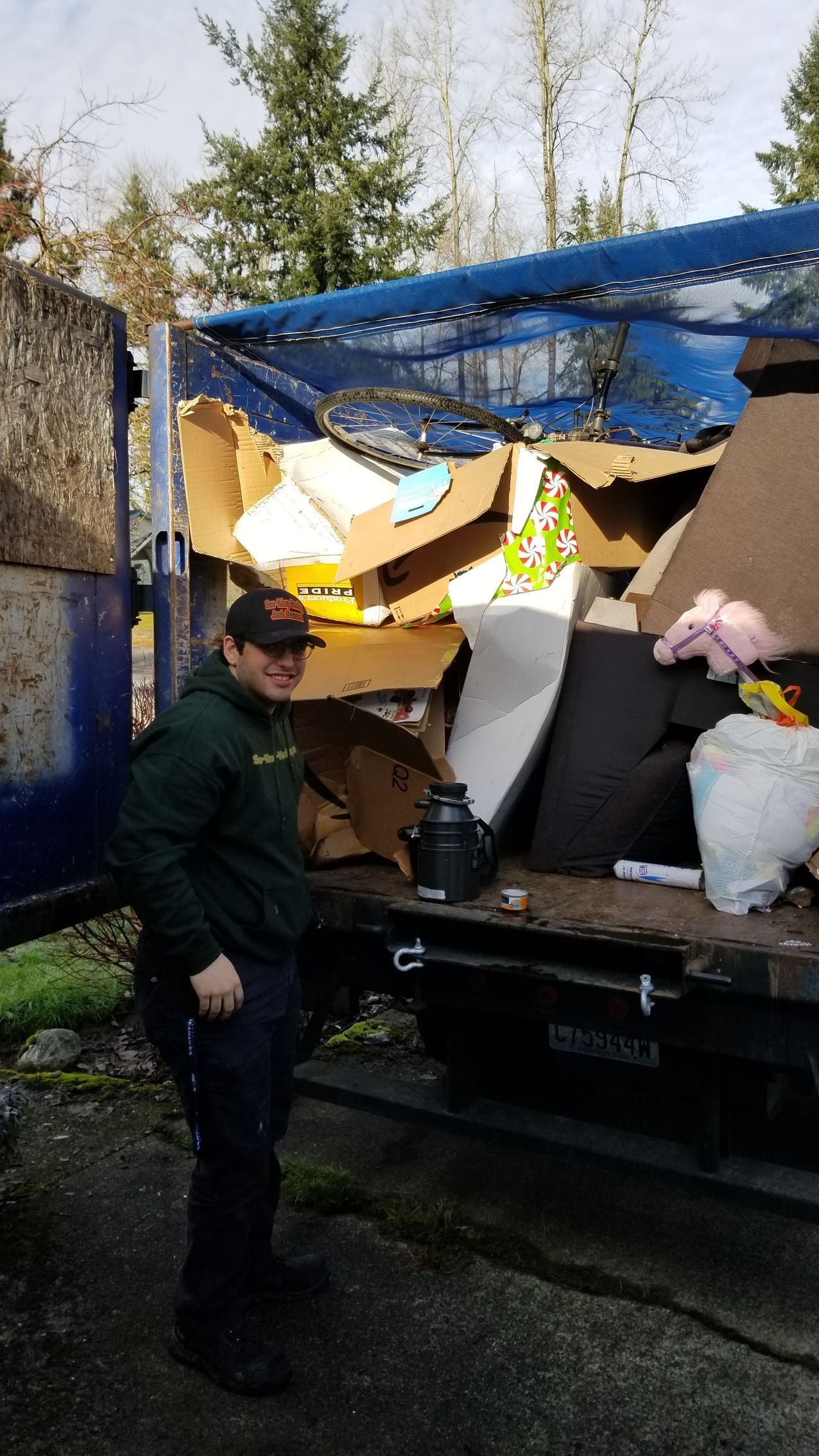 A person wearing a dark hoodie and cap stands smiling next to a truck bed filled with assorted cardboard and trash.