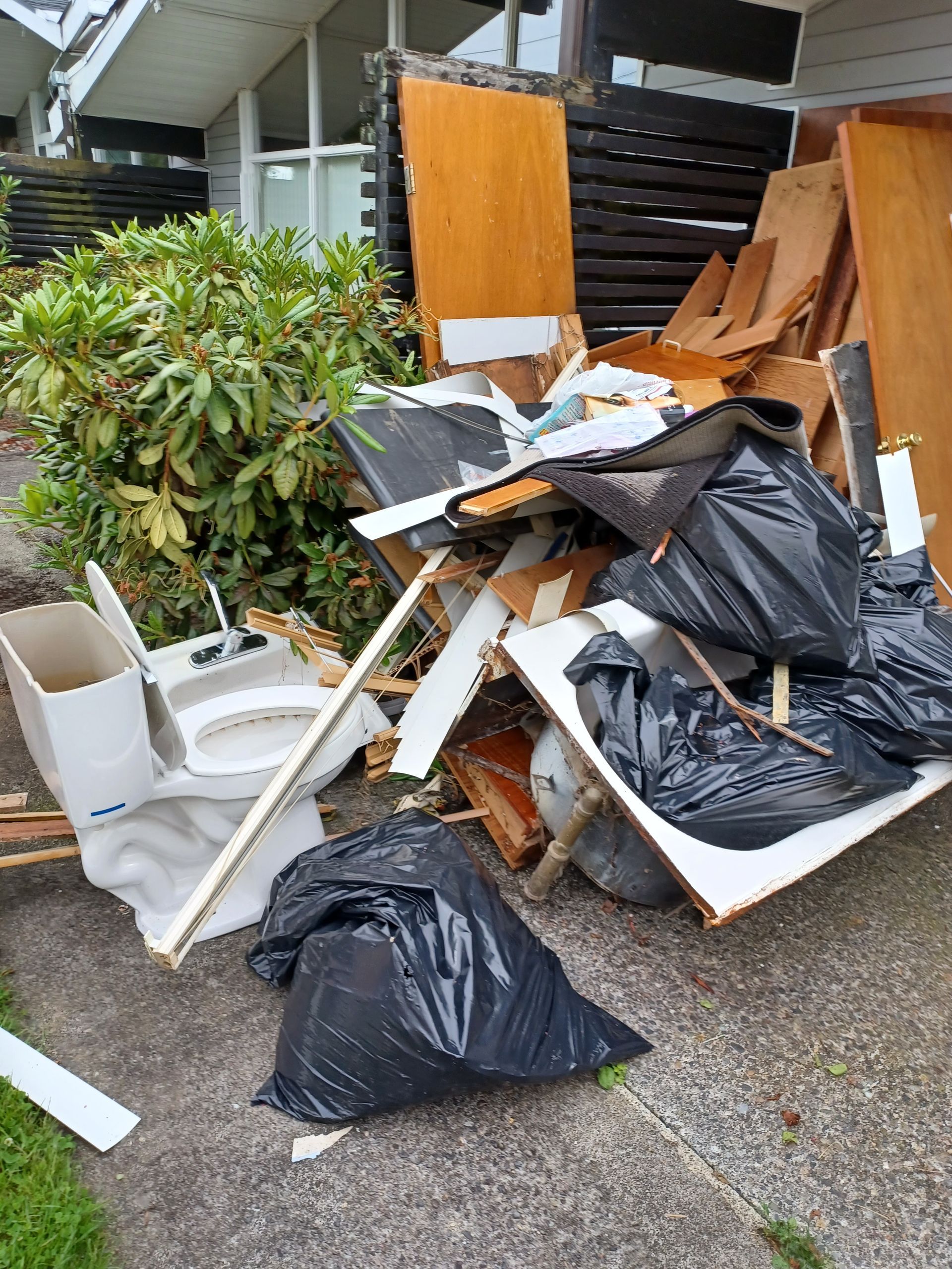A pile of residential waste, including a toilet, wood scraps, and black trash bags, sits outside a house.