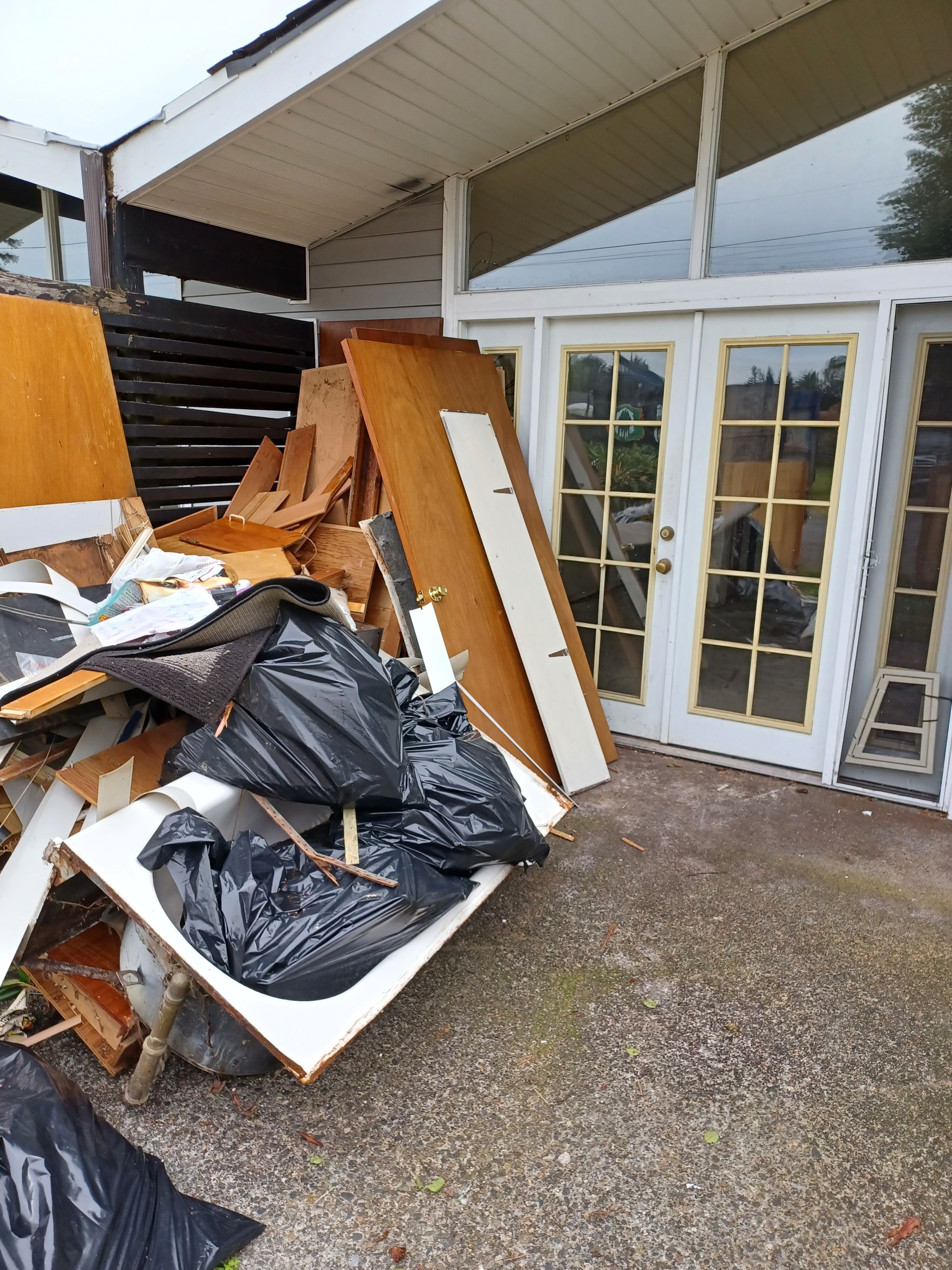 A pile of debris, wooden boards, and black trash bags sits on a patio in front of white-framed glass doors.