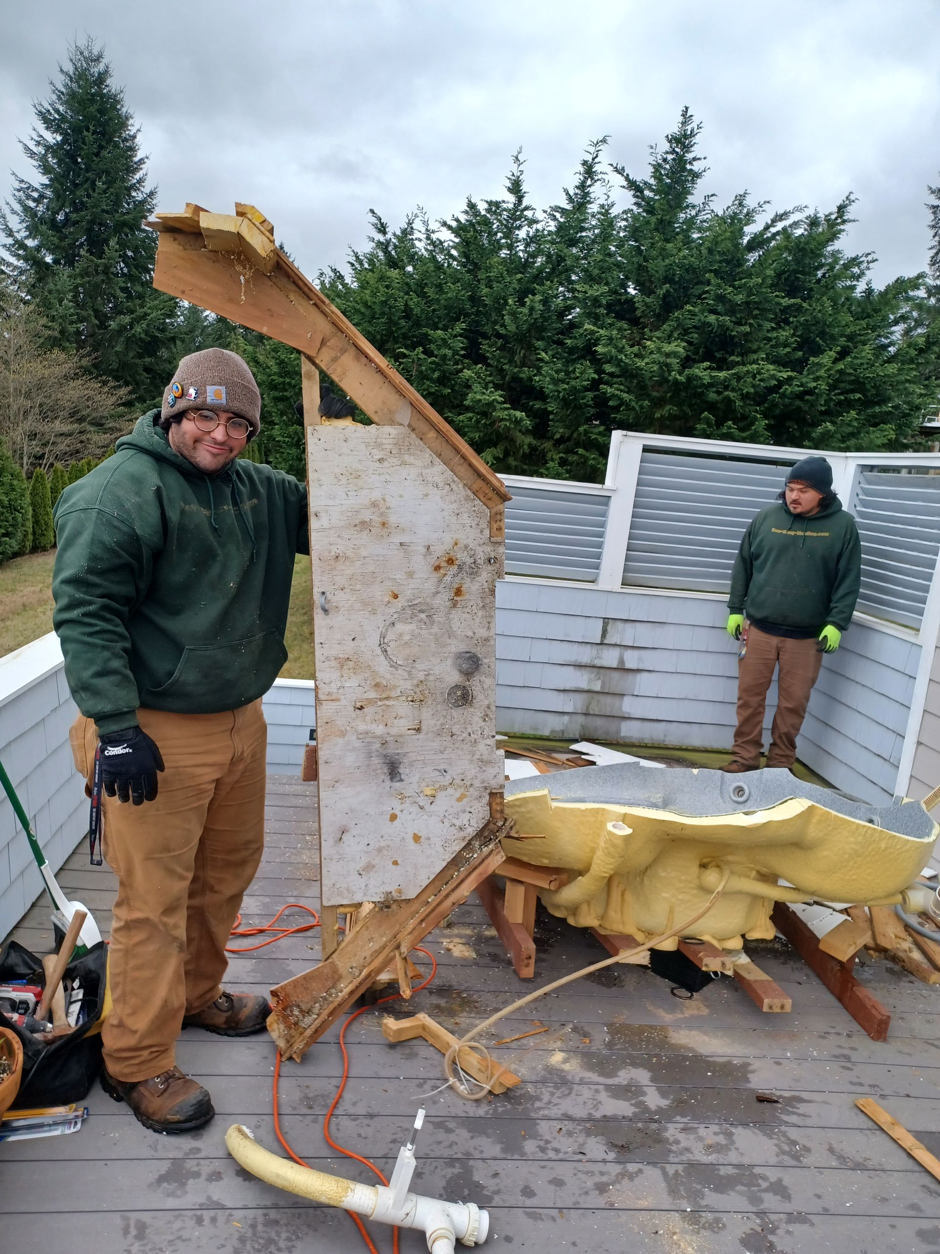 Two people in work clothes stand on a wooden deck next to large pieces of dismantled light-colored construction material.