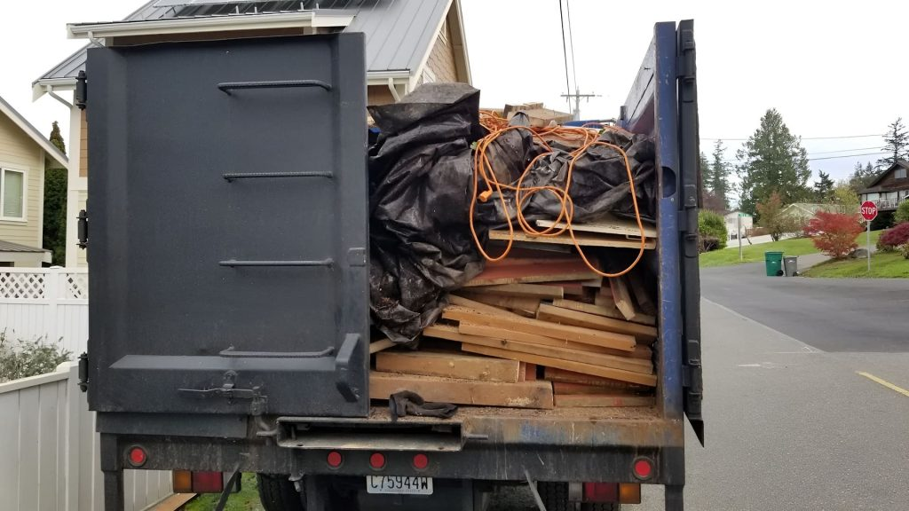 Rear view of a dark blue dump truck filled with scrap wood and a black tarp parked on a residential street.