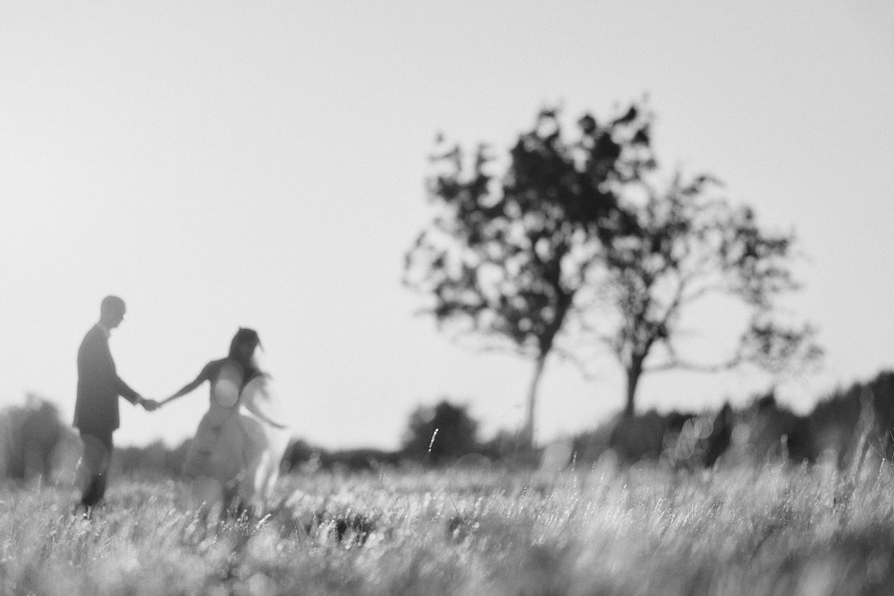 Couple holding hands, walking in a field towards a blurred tree, black and white photo.