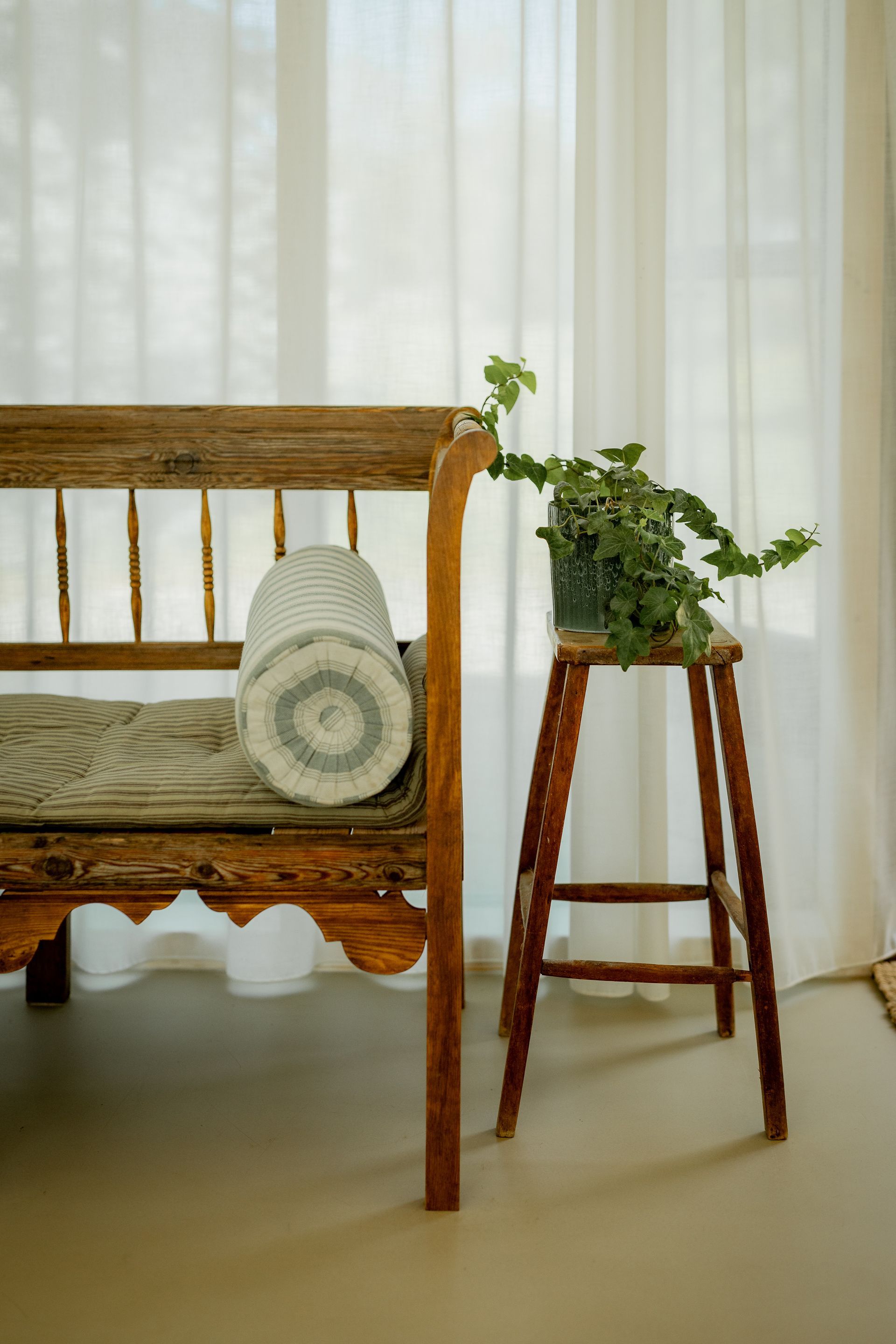 Wooden bench and stool with decorative pillow and potted plant in front of sheer curtains.