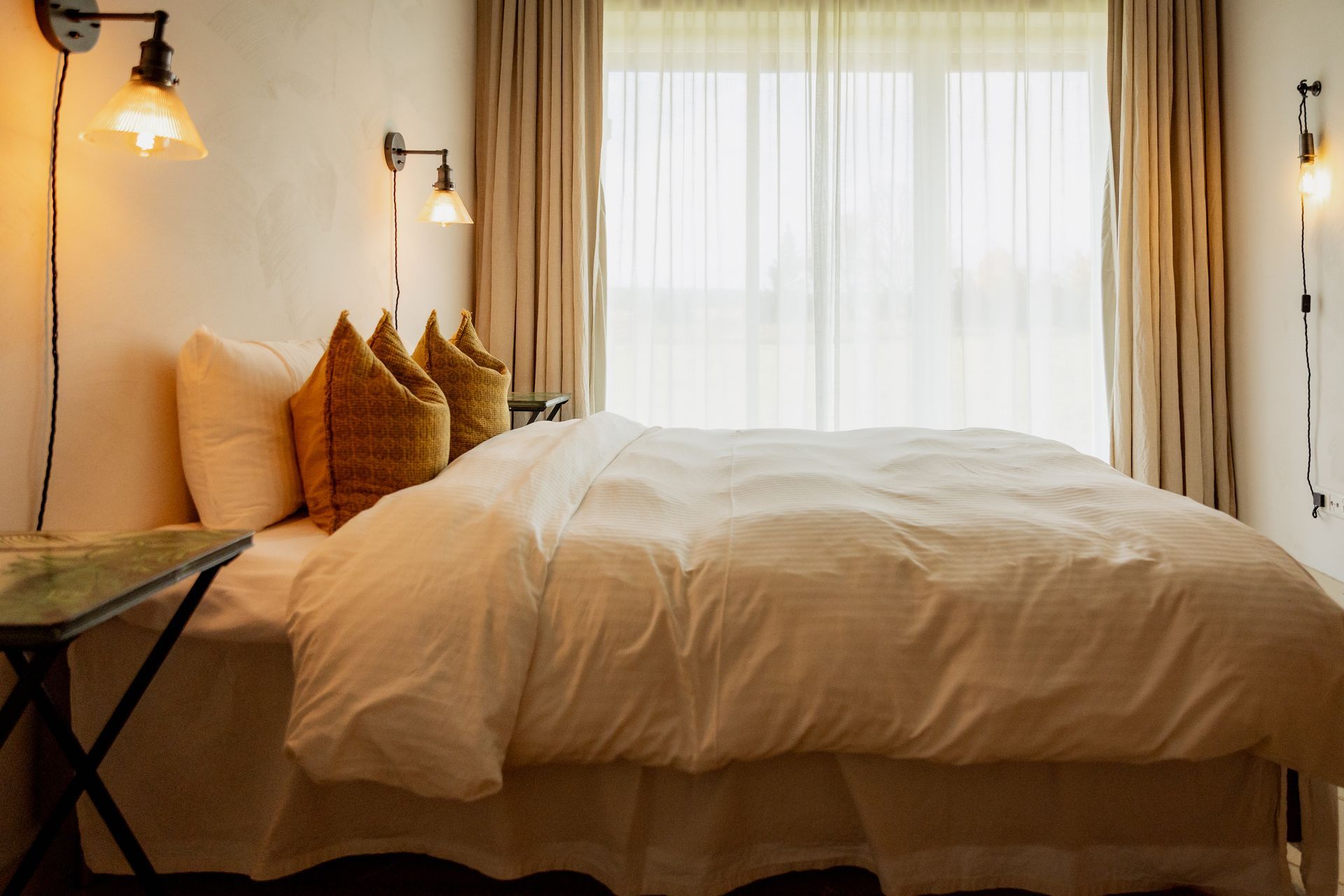 Bedroom with a large bed, tan walls, and a window with sheer curtains. Three sconces and a small table.