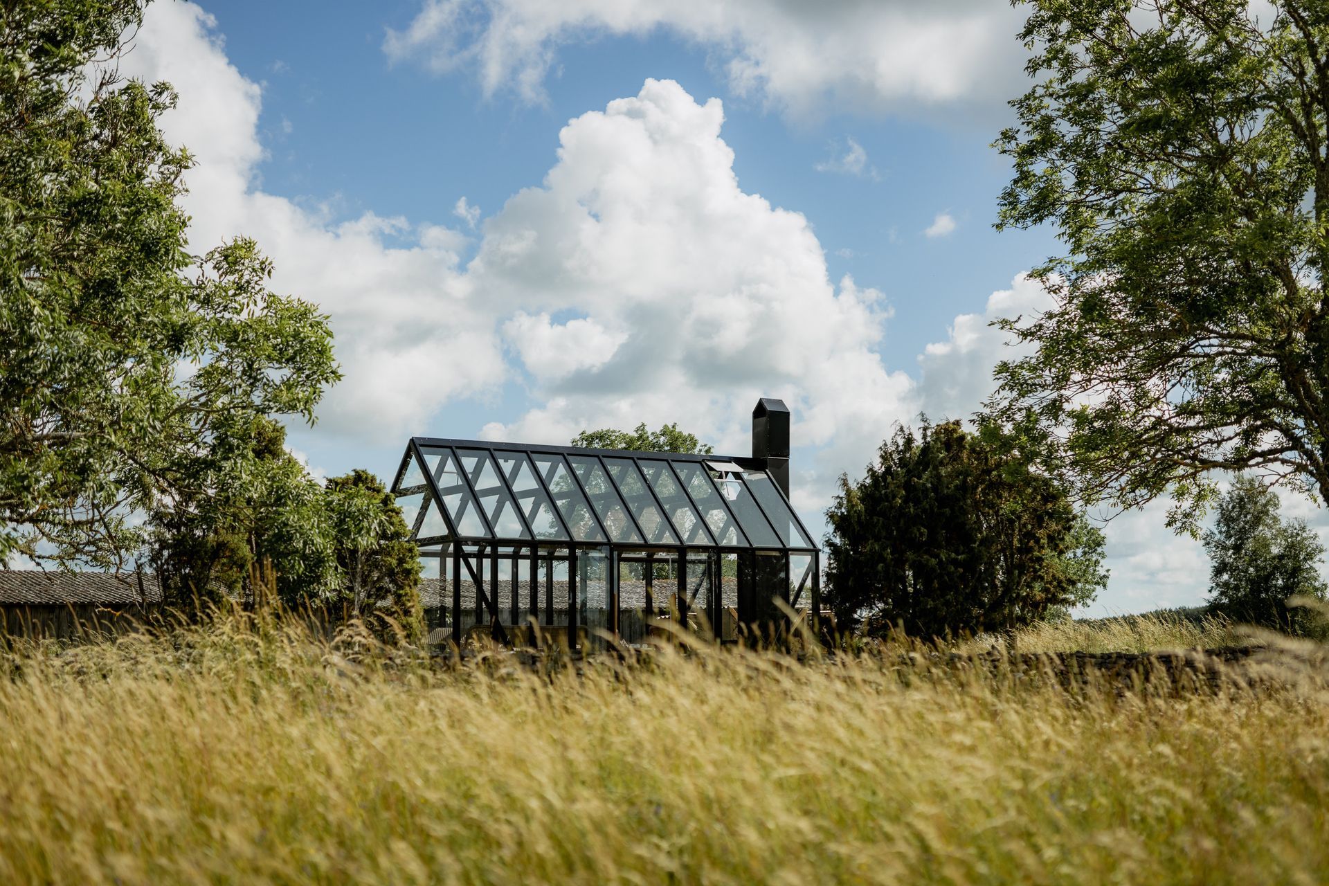 Glass greenhouse with black frame in a grassy field, under a cloudy blue sky.