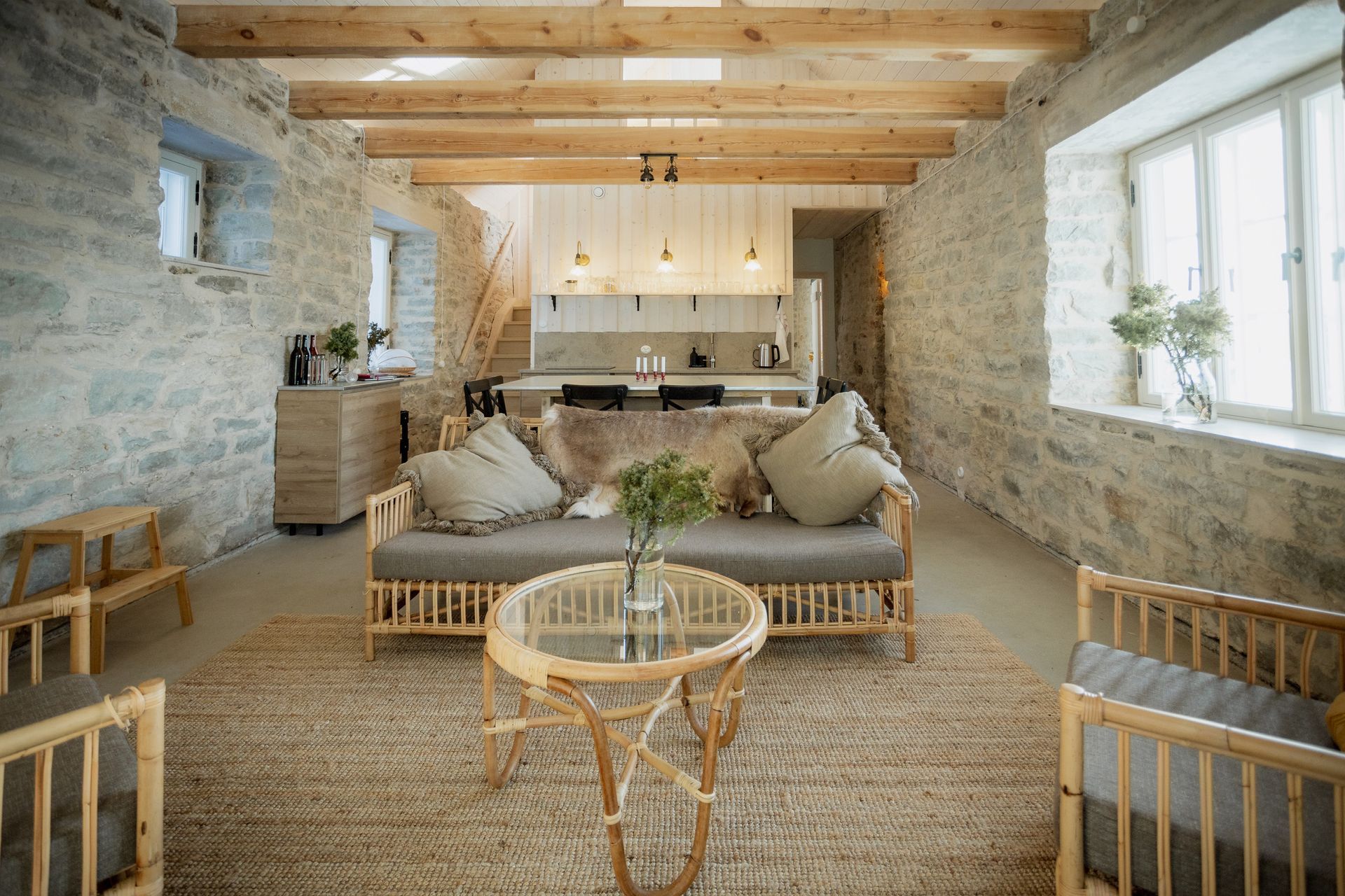 Living room with stone walls, wooden beams, rattan furniture, and an open kitchen.