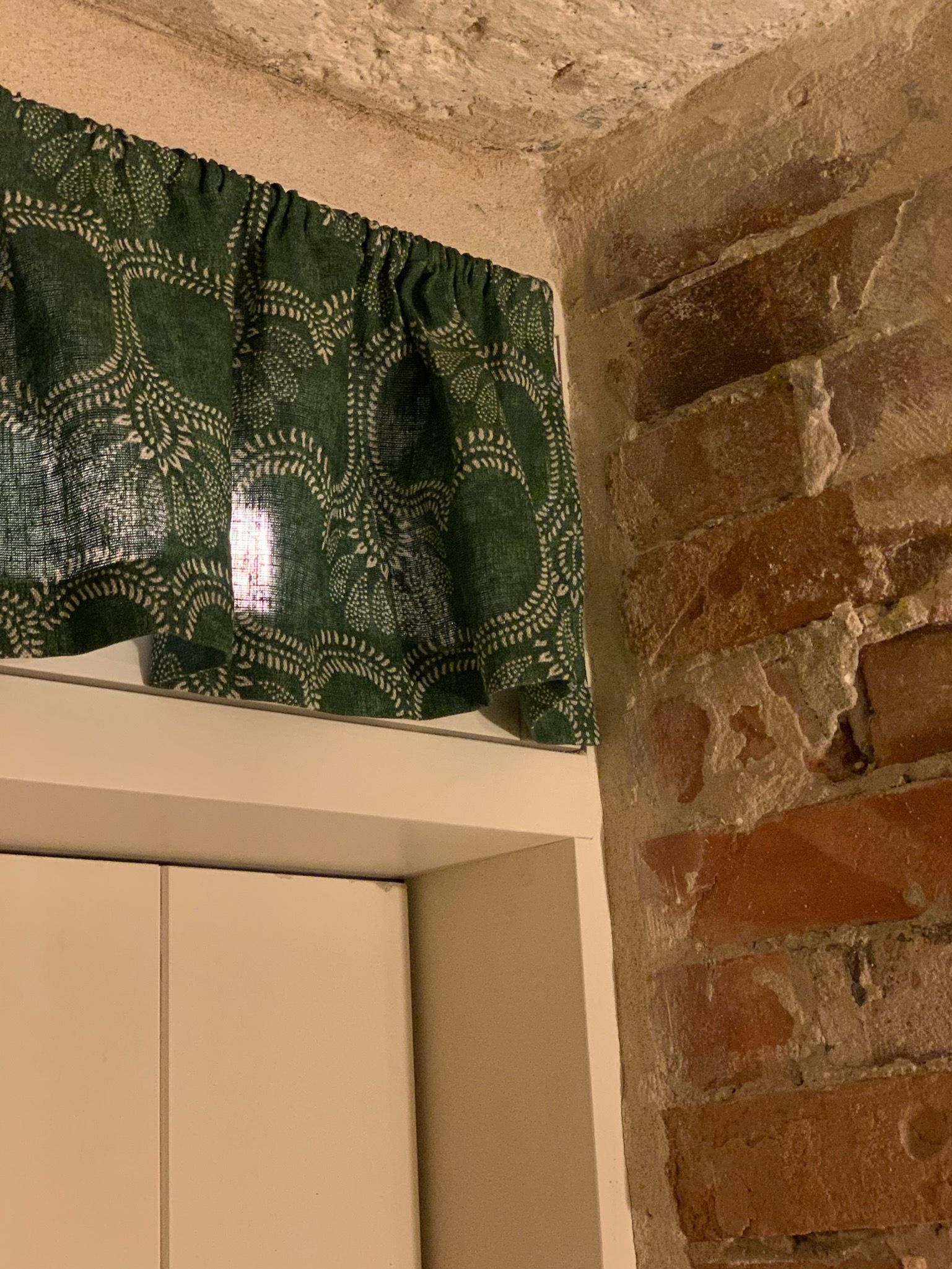 Green patterned curtain above a white door and exposed brick wall corner.