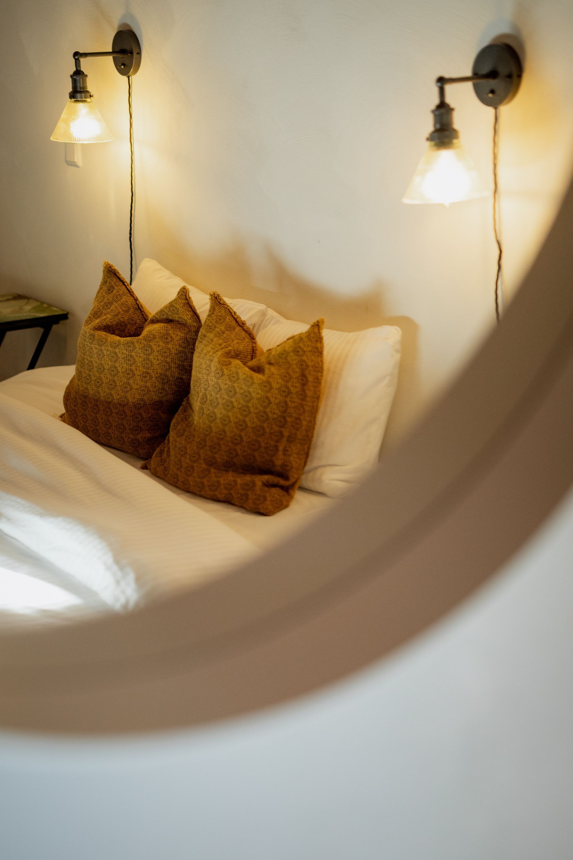Bed with pillows and sconce lighting, seen through a blurred circular mirror.