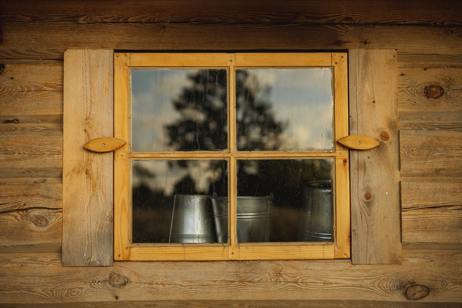 Wooden window in a rustic cabin, reflecting trees and metal buckets.