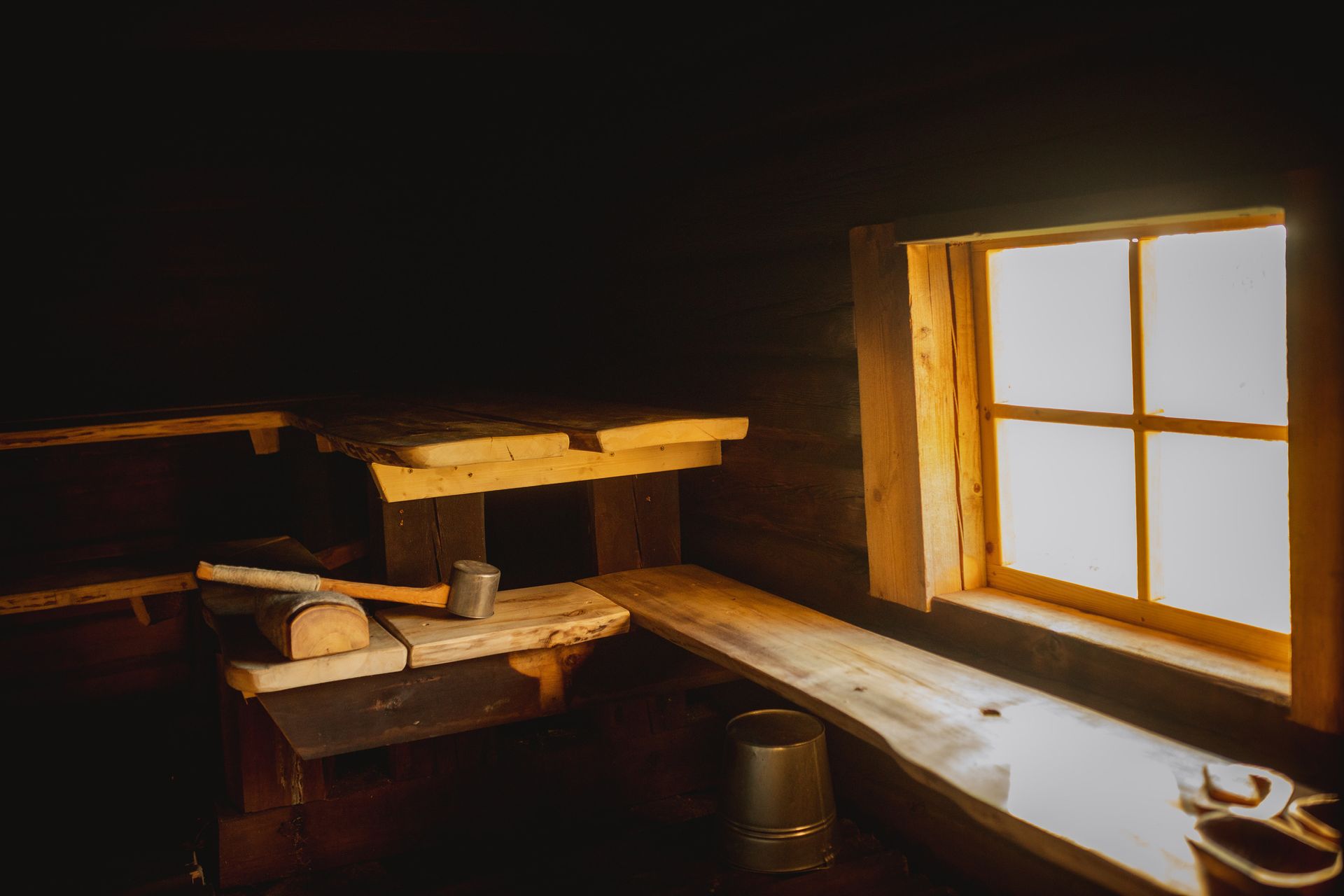 Interior of a wooden sauna lit by sunlight from a window, featuring wooden benches and a small metal bucket.