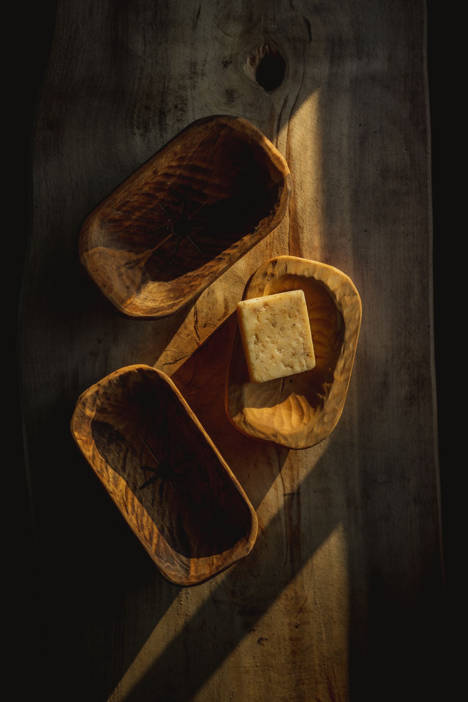 Wooden bowls on a wood surface, one with a square of cheese, lit by sunlight.
