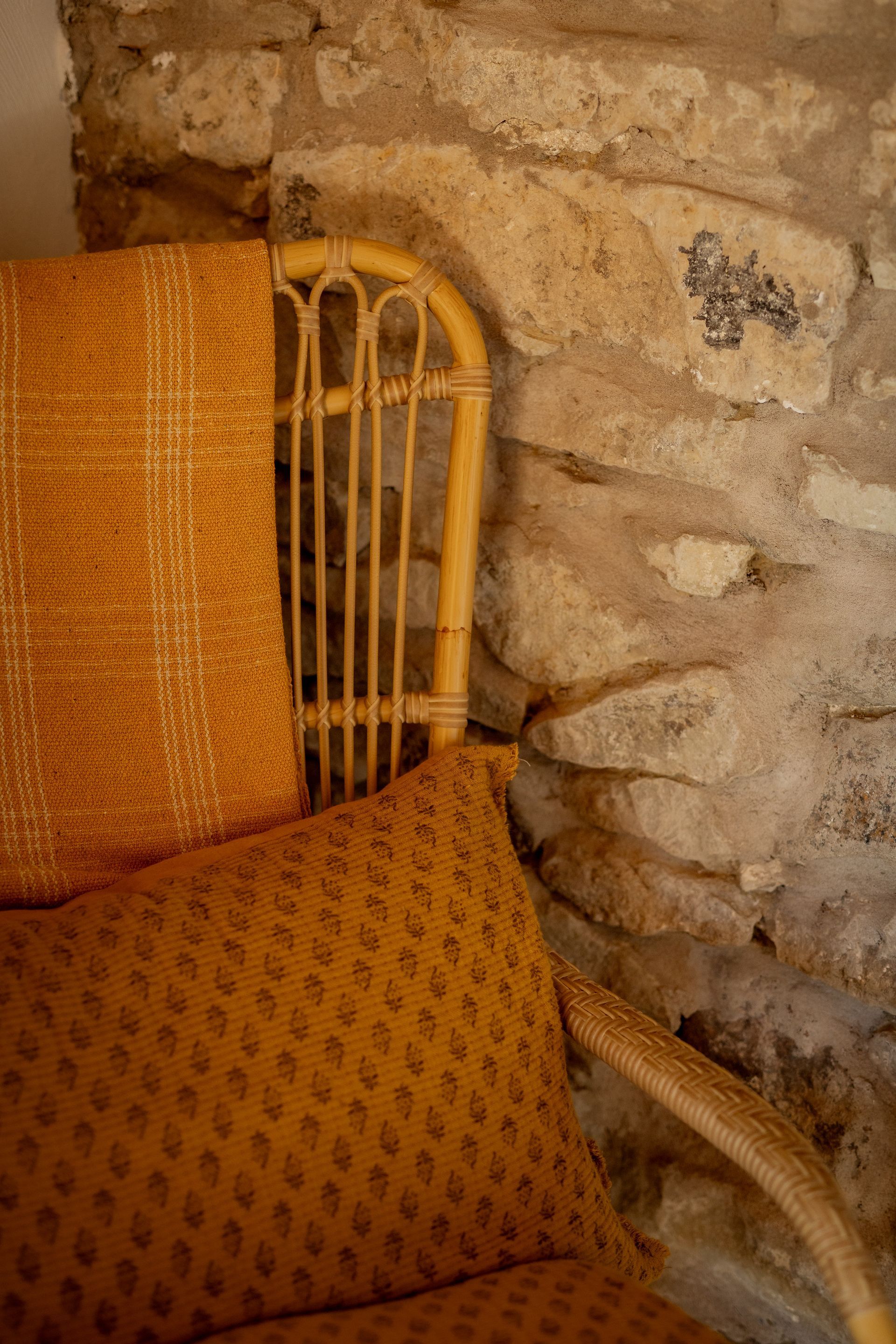 Wicker chair with orange cushions and a blanket, positioned next to a rough stone wall.