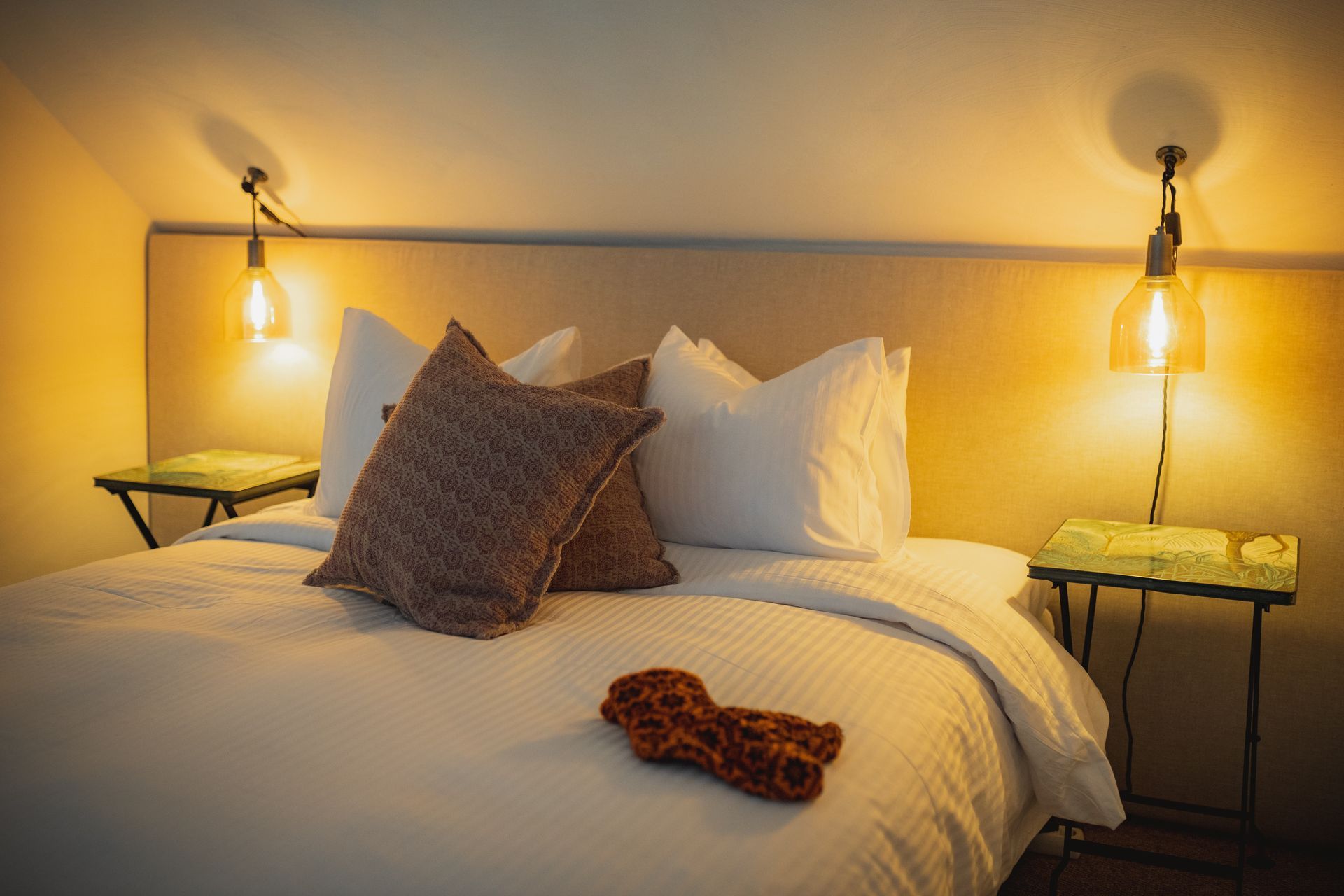 Cozy bedroom with bed, pillows, lamps, and small side tables, warm lighting.